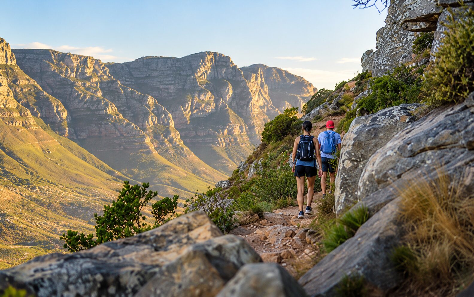 Two hikers walking along a rocky mountain trail, with sweeping views of the mountains surrounding Cape Town.